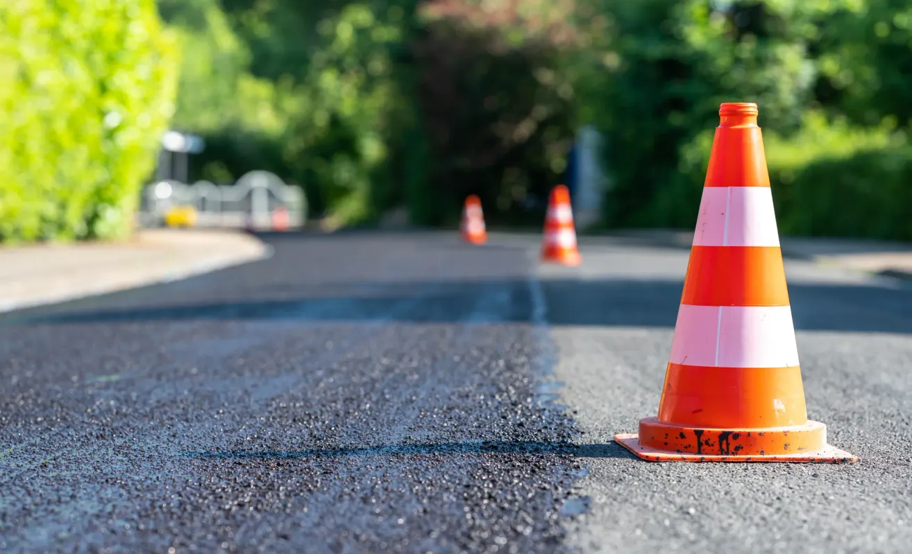 A road with fresh asphalt and orange traffic cones placed to guide traffic. Trees and greenery are visible in the background.