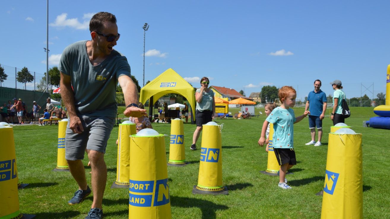 A man throws a ball at a yellow inflatable target on a grassy field. A woman and a child watch nearby. In the background, there is a yellow tent and a yellow building.
