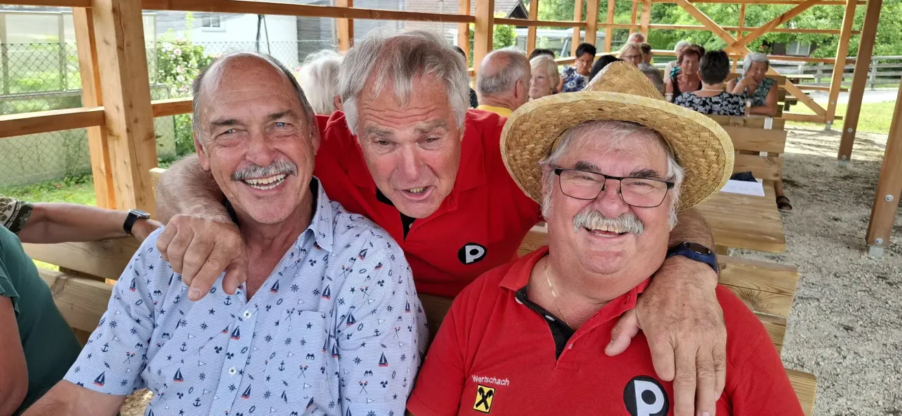 Three older men are smiling and posing for a photo. The man on the left wears a light blue shirt with a nautical pattern. The man in the middle, wearing a red shirt, has his arm around the man on the right, who is wearing a straw hat and glasses.
