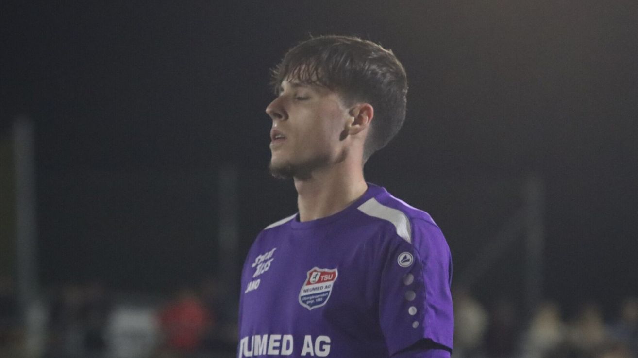 Ein Fußballspieler mit einem lila Trikot, trägt ein Logo auf der Brust und ein Sponsorenlogo am Ärmel.