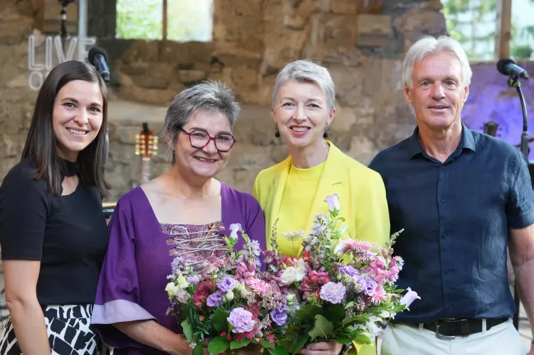 Four individuals, two women and two men, are standing together and smiling, holding bouquets of flowers. The woman on the left wears glasses and a purple blouse, while the woman in the center wears a yellow blazer. The man on the right wears a blue polo.