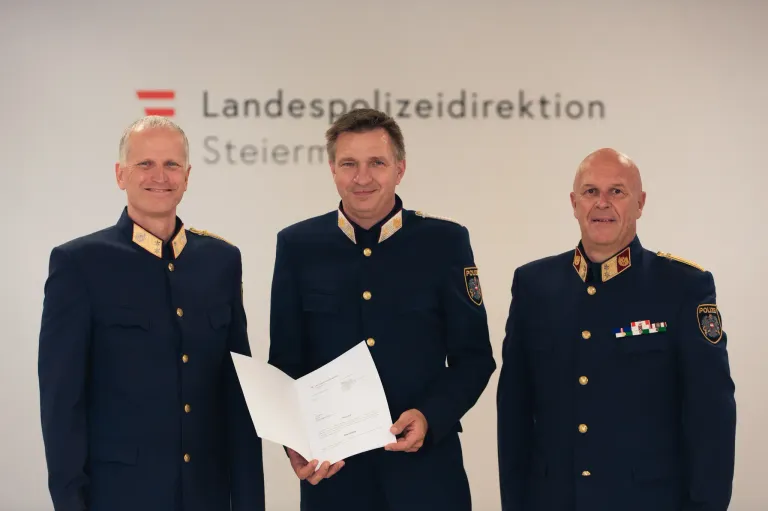 Drei Männer in Marineuniformen stehen vor einer weißen Wand mit einem Schild, das 'Landespolizeiinspektion Steiermark' besagt. Der Mann in der Mitte hält ein Papier, während die anderen lächeln.