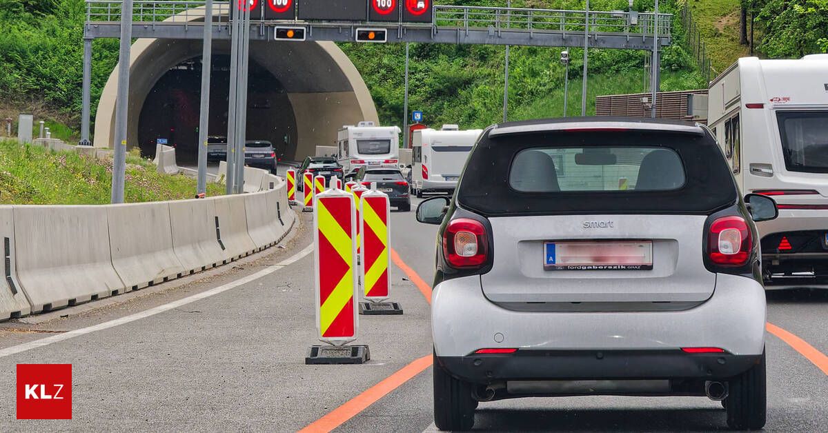 A Smart car is stopped at a traffic barrier on a road with cars and trucks in the distance. The road has a tunnel and a bridge above.