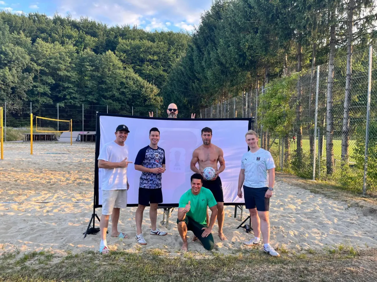 Five men pose for a photo on a sandy beach with a large banner behind them. One man kneels in the foreground, holding a soccer ball.