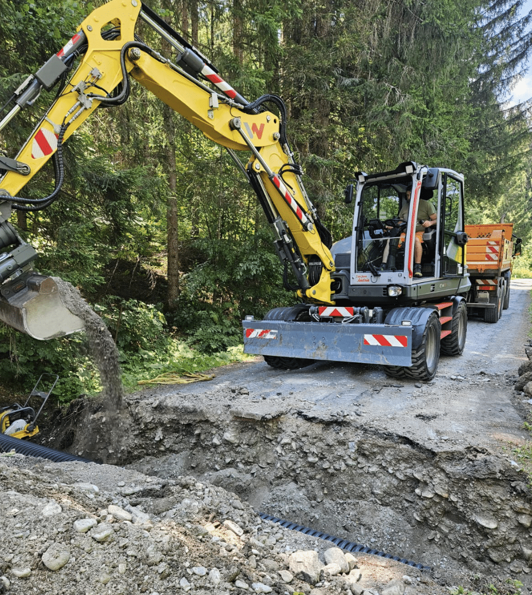 A yellow backhoe loader is dumping soil into a hole on the side of a road. A person is operating the machine.
