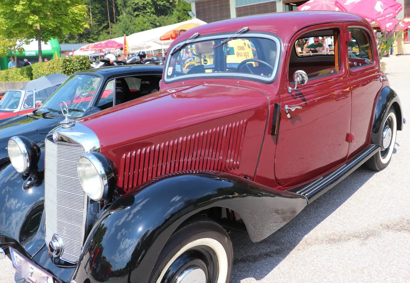 A red vintage car with a shiny black bumper and chrome accents is parked in an outdoor setting. The car's front end features a classic design with a prominent grille and round headlights.