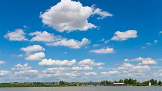 Eine friedliche Landschaft mit einem Gewässer unter einem blauen Himmel voller flauschiger Wolken und einem entfernten Ufer mit Bäumen und Gebäuden.