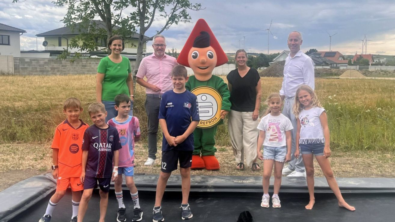 A group of adults and children pose for a photo with a mascot in a field. The mascot wears a red hat and a green costume with a logo. The children wear sports jerseys.
