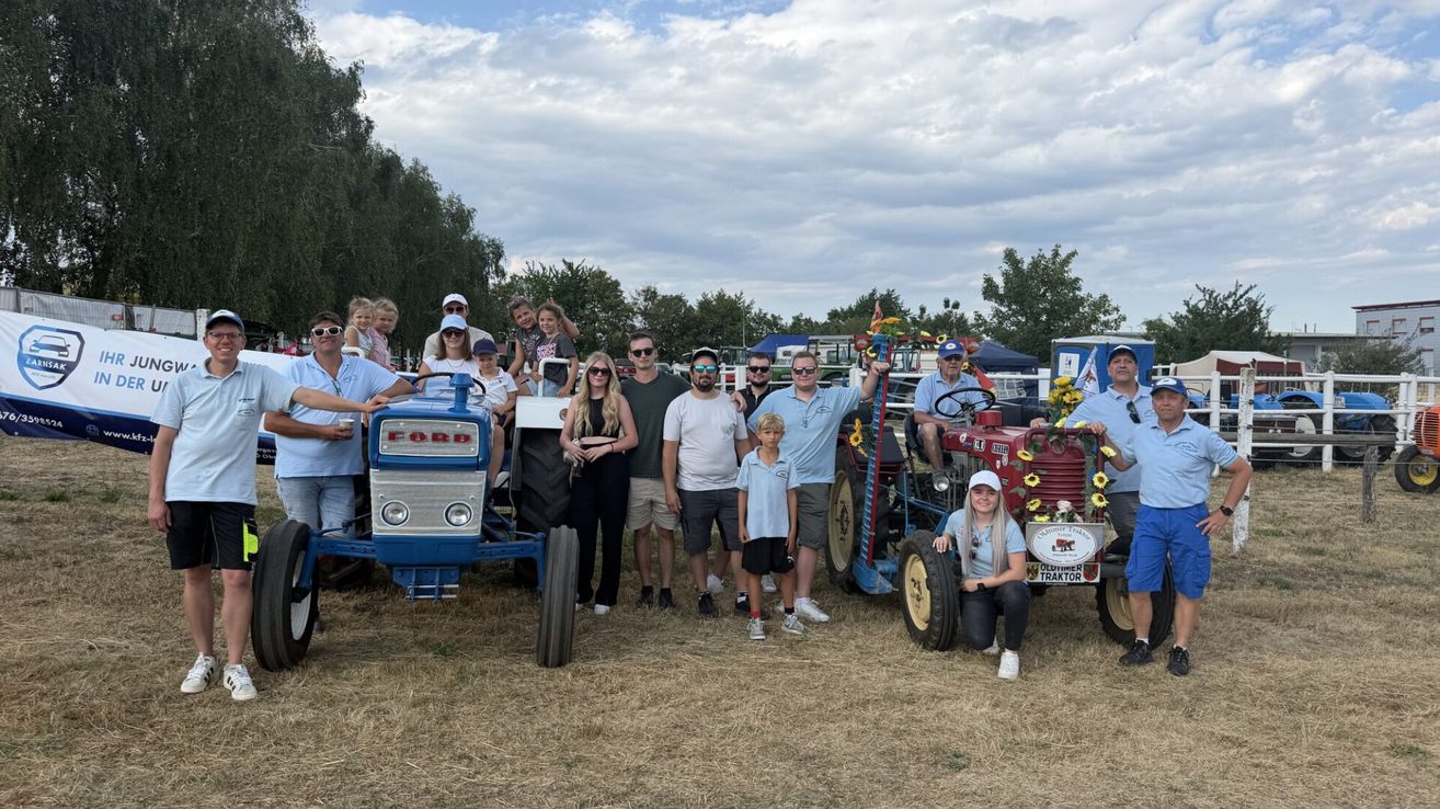 A group of people are posing for a photo in front of two vintage tractors. They are wearing casual clothing and some are wearing sunglasses. The tractors are blue and red.