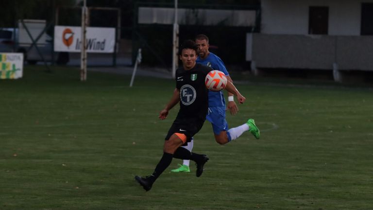 A soccer player in black chases a ball while another player in blue runs behind him on a field.