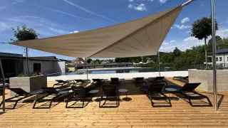 Eine Außenlounge mit einem großen beigen Baldachin, mehreren schwarzen Liegestühlen und kleinen Tischen, vor dem Hintergrund eines Schwimmbeckens und eines blauen Himmels mit Wolken.