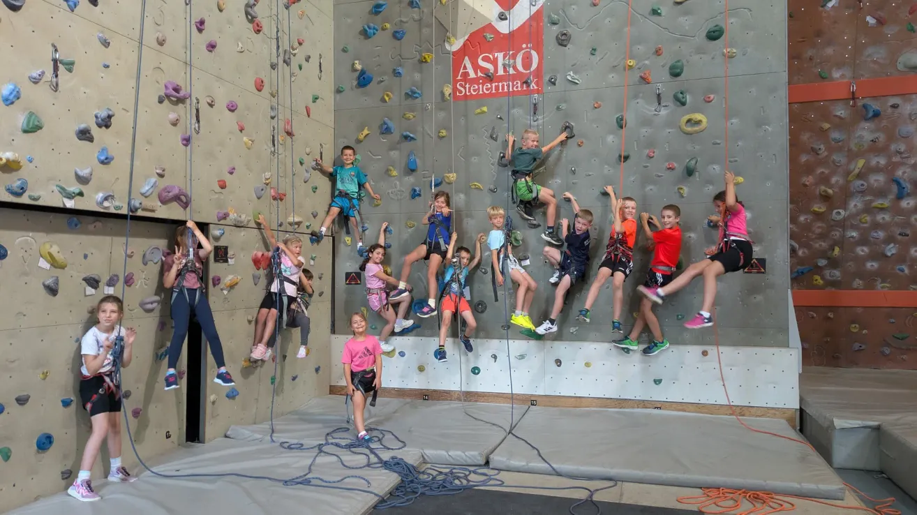 A group of children are climbing a rock wall with colorful holds. Some are mid-climb while others are at the bottom. A sign reading 'ASKO Steiermark' is visible on the wall.
