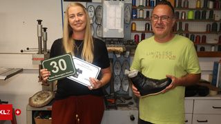 A woman and a man stand in a shoe repair shop, the woman holding a sign with the number 30 and a certificate, the man holding a black shoe.