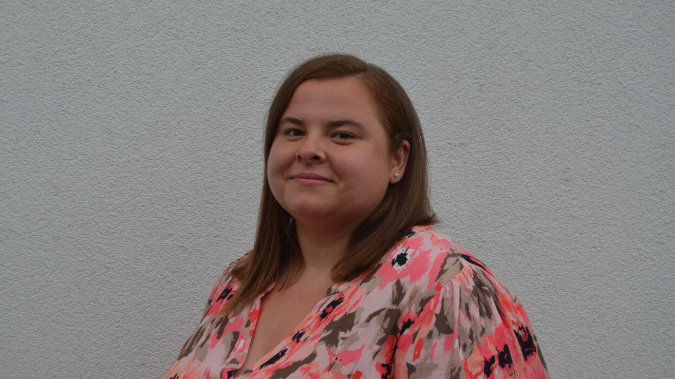 A woman with brown hair wearing a pink floral blouse is smiling and standing against a white wall.