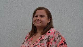 A woman with brown hair wearing a pink floral blouse is smiling and standing against a white wall.