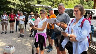 Eine Gruppe von Menschen, wahrscheinlich auf einer Wanderung, versammelt sich und liest einen Prospekt. Sie sind in lässiger Outdoor-Kleidung gekleidet, einige tragen Brillen und Hüte. Ein Van ist hinter ihnen geparkt.