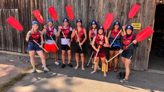 Eine Gruppe von Menschen, die in Schwimmwesten und Helmen gekleidet sind, Paddel halten und vor einer Holzwand für ein Foto posieren.