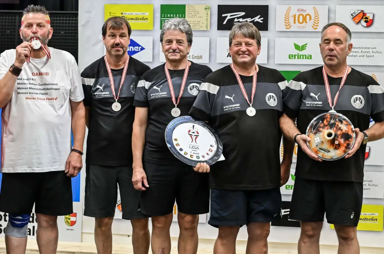 Four men in matching black and gray sports uniforms with medals around their necks stand together, smiling, and holding a plaque and a trophy. They are in front of a wall with various logos and text.