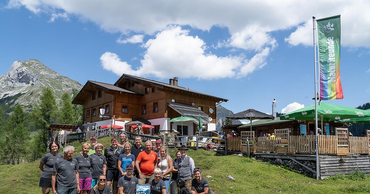 A group of people stands in front of a wooden building with a mountain in the background. They are all wearing matching t-shirts and some are holding a trophy. The building has a green umbrella and a fence.
