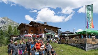 A group of people stands in front of a wooden building with a mountain in the background. They are all wearing matching t-shirts and some are holding a trophy. The building has a green umbrella and a fence.
