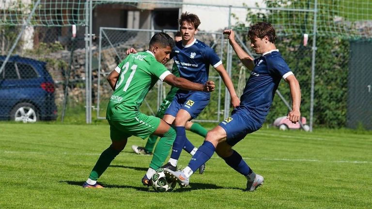 Drei Fußballspieler sind in einem Spiel auf einem Feld engagiert. Der Spieler in Grün versucht, den Ball vom Spieler in Blau zu stehlen. Ein anderer Spieler in Blau beobachtet sie.