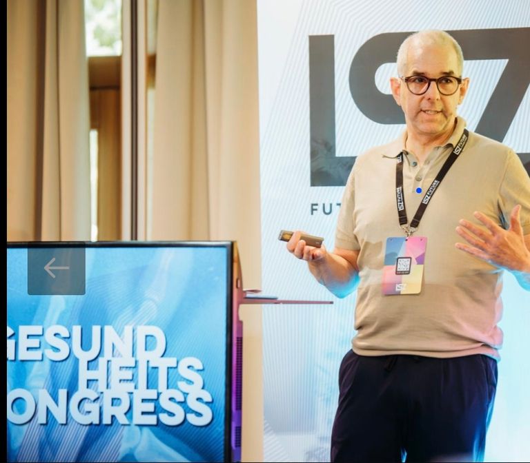Ein Mann mit Brille und beigem Poloshirt steht vor einem Bildschirm mit der Aufschrift 'Gesundheits Kongress'. Er hält ein kleines elektronisches Gerät in der Hand und scheint eine Präsentation zu halten.