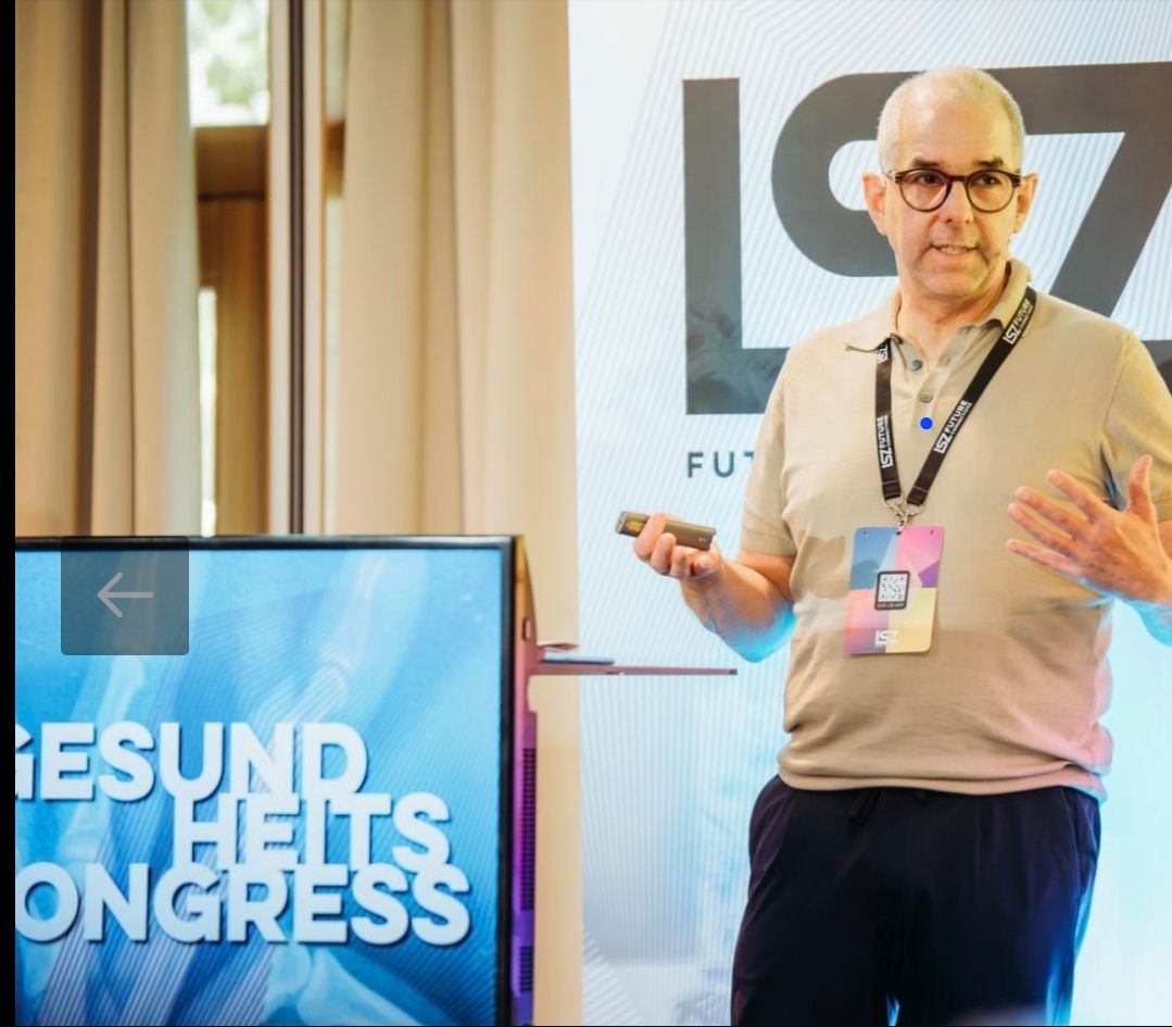 Ein Mann mit Brille und beigem Poloshirt steht vor einem Bildschirm mit der Aufschrift 'Gesundheits Kongress'. Er hält ein kleines elektronisches Gerät in der Hand und scheint eine Präsentation zu halten.