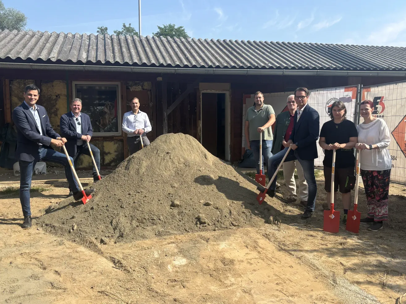 A group of people in business attire are gathered around a pile of dirt, holding shovels, and posing for a photo. Behind them is a building with a metal roof.