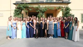 A group of women in evening gowns pose for a photo in front of a building with green shutters and climbing plants.