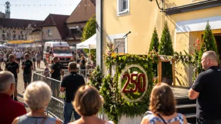 Eine Straßenszene mit einem dekorierten Podium mit der Nummer 250, einem Mikrofon und umstehenden Leuten. Ein roter Krankenwagen ist in der Nähe geparkt.