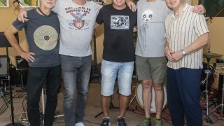 Five men stand together in a room, smiling and posing for a photo. They are dressed casually and appear to be friends. Behind them, there is a drum set, a microphone, and some guitars.