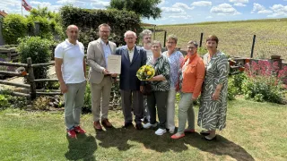A group of people stand in a garden, smiling and posing for a photo. A man holds a certificate, and a woman holds a bouquet of flowers. Behind them, there is a fence and a field.