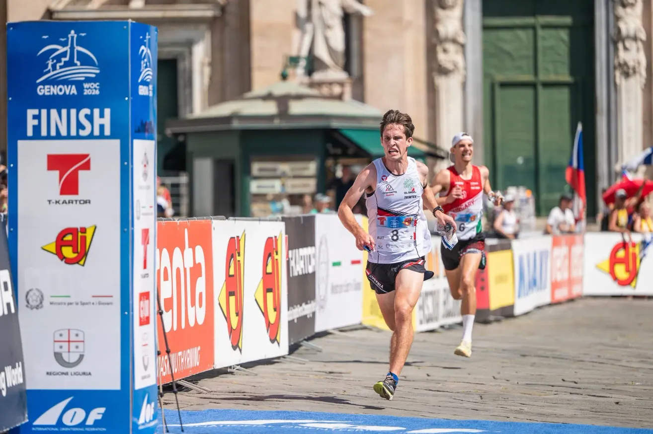 Ein Läufer mit der Nummer 8 auf seinem Trikot sprintet bei einem Marathon, dicht gefolgt von einem anderen Läufer. Sie laufen auf einer Kopfsteinpflasterstraße mit Bannern und einer Statue im Hintergrund.