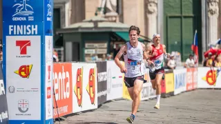 Ein Läufer mit der Nummer 8 auf seinem Trikot sprintet bei einem Marathon, dicht gefolgt von einem anderen Läufer. Sie laufen auf einer Kopfsteinpflasterstraße mit Bannern und einer Statue im Hintergrund.