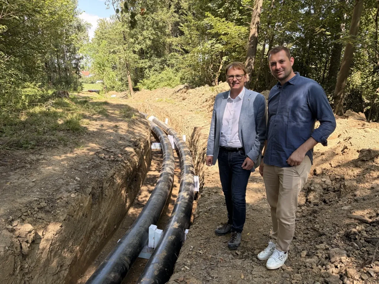 Two men stand next to each other in a construction site with large pipes buried in the ground and trees in the background.