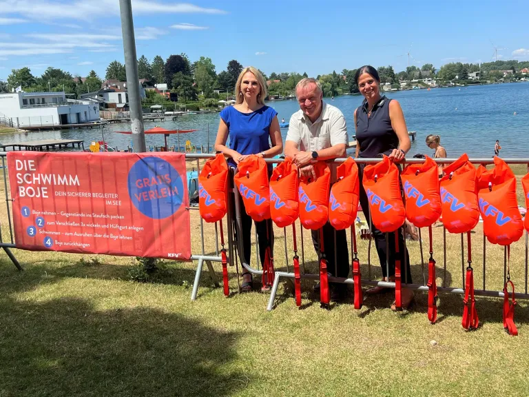 Three people stand behind a barrier with orange life vests hanging from it. A banner is visible in the background.