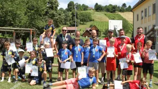 A group of young athletes and coaches pose for a photo on a grassy field, holding certificates and medals, with a scenic backdrop of trees and houses.