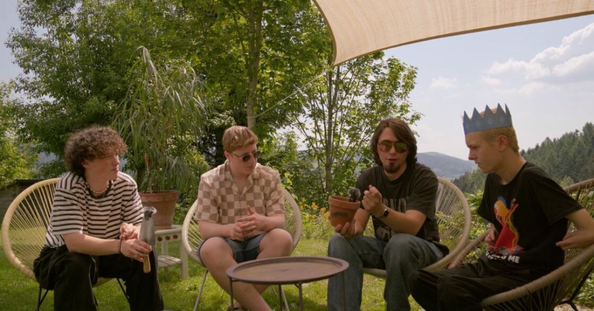 Four men sit in chairs in a garden, one holds a potted plant, another gestures with a tool. A table and a tree are in the background.