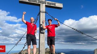 Two men in red shirts pose at a mountain peak, one with a fist raised, both holding onto chains attached to a wooden cross.
