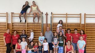 A group of people, including children and adults, are posing for a photo in a gym. Two women are on the horizontal bars, and the rest are standing and sitting on the floor.