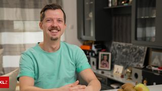 A man in a mint green t-shirt is smiling and sitting in a room with a blurred background. There are picture frames and a globe on a desk behind him.