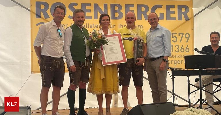 Five individuals stand on a stage, smiling for a photo. A woman in the center holds a bouquet and a certificate. The man on the right wears a watch and holds a trophy. A banner with the word 'Rosenberger' is behind them.