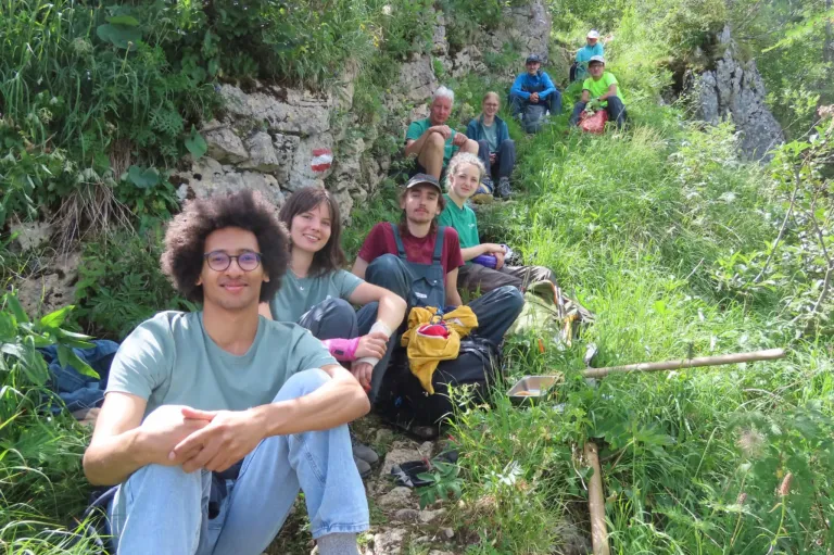 A group of people are sitting on the ground in a grassy area, some wearing hats and glasses, with a stone wall in the background.
