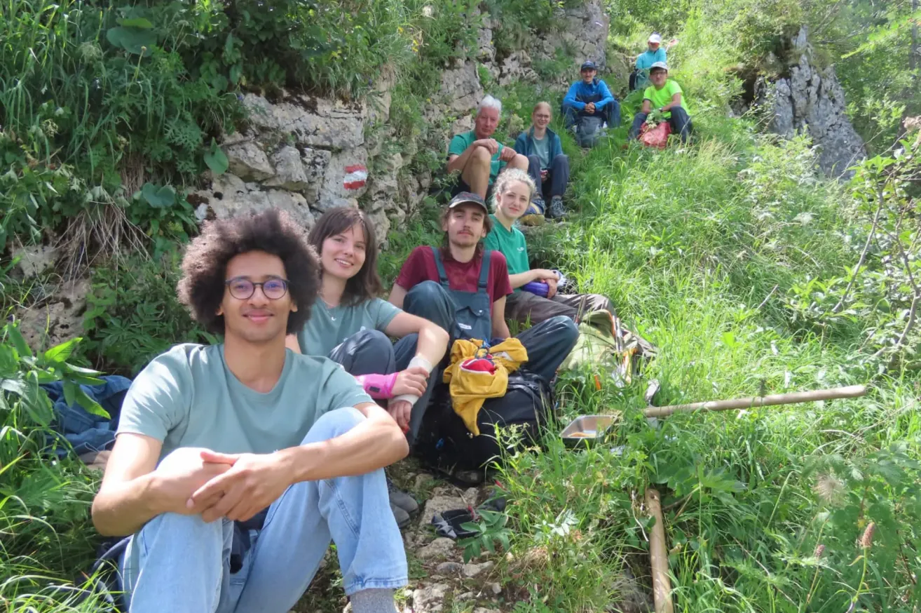 A group of people are sitting on the ground in a grassy area, some wearing hats and glasses, with a stone wall in the background.
