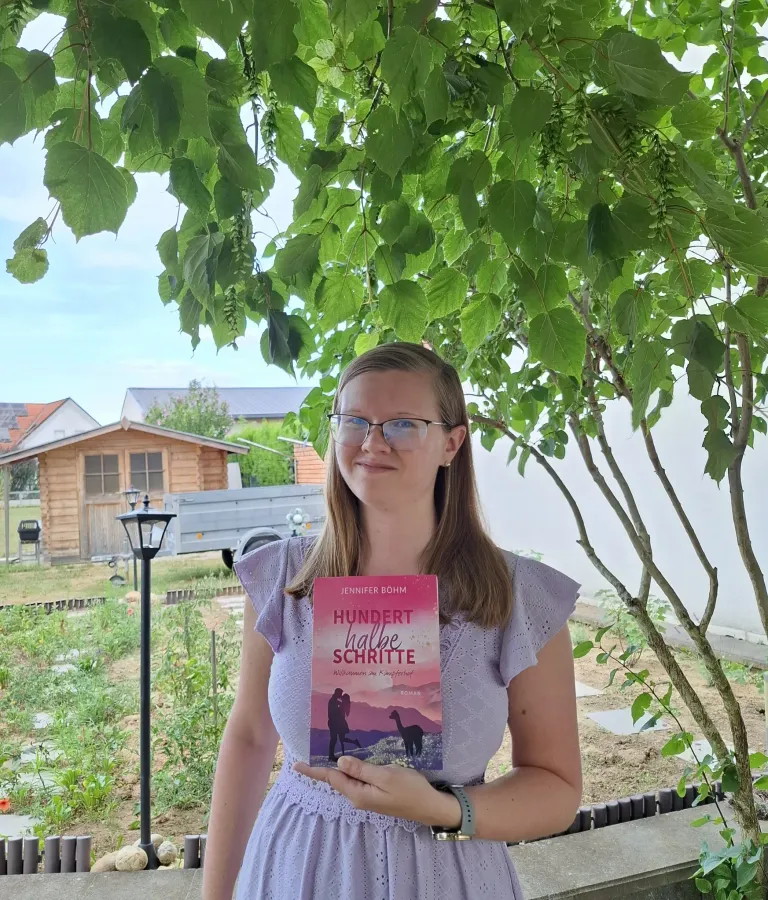 Eine Frau in einem lila Kleid hält ein Buch mit dem Titel 'Hundert halbe Schritte' in einem Garten unter einem Baum.