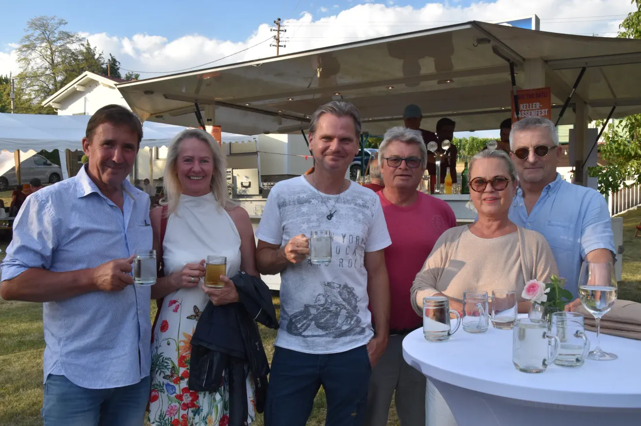 A group of people, including a man in a white shirt with a graphic design, are standing together and holding glasses of beer. They are smiling and posing for a photo.