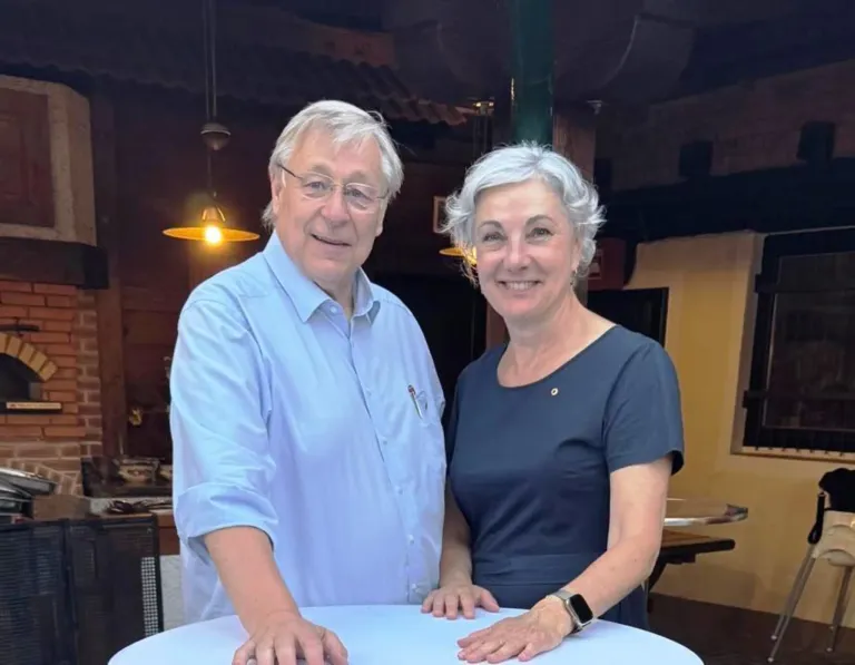 An elderly man and woman stand together at a table, smiling for a photo. The man wears glasses and a light blue shirt, while the woman wears a dark blue top. Behind them is a warm-lit room with a hanging lamp.