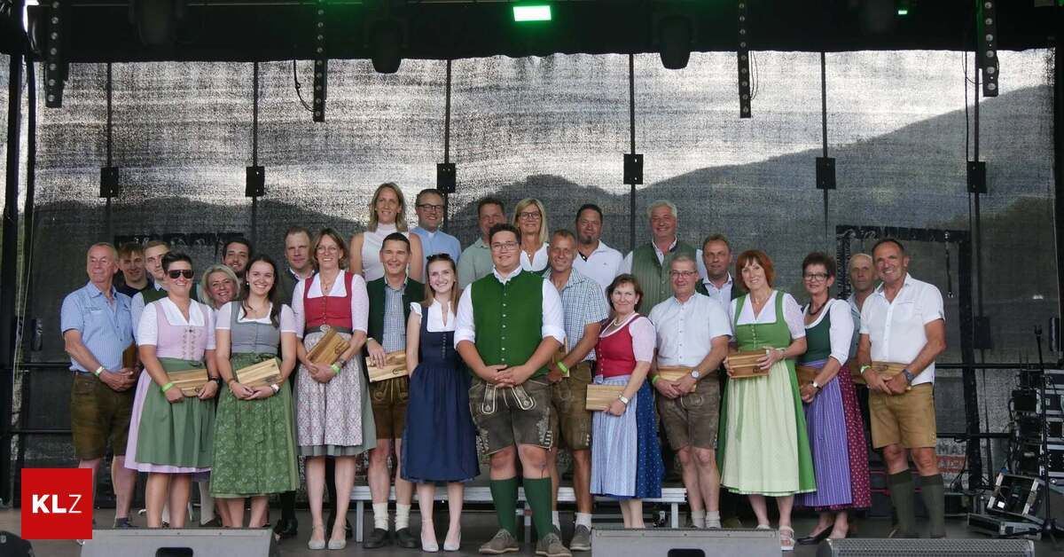A group of people dressed in traditional German attire are posing for a photo on a stage with a backdrop showing mountains. They hold wooden awards and appear to be celebrating an event.