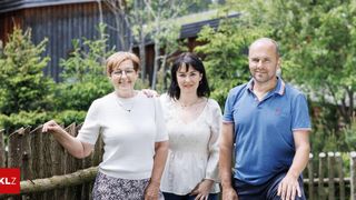 Three people are standing together in a garden. The woman on the left is smiling and holding a wooden fence. The woman in the middle is looking at the camera. The man on the right is smiling.