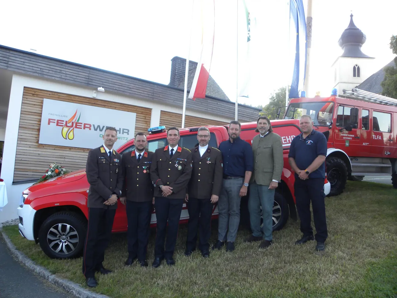 A group of men in uniforms and suits stand in front of a red fire truck with a banner behind them.